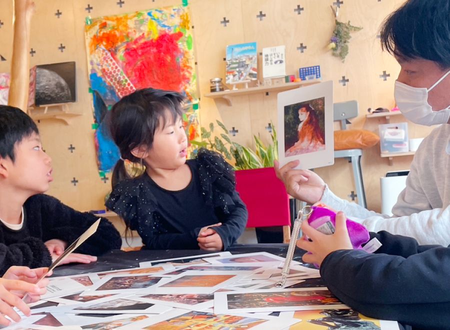 机に座ってパパンダ先生が持っているアート作品を観察している2人の子供。机の上にはたくさんのアート作品が広げられている