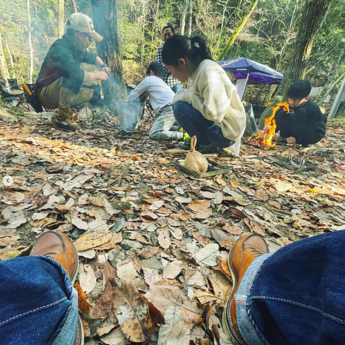 山の中のたくさんの落ち葉の前で焚き火をしている子供達と、それを見守る大人